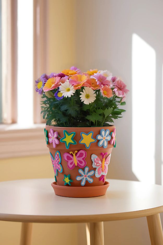 A terracotta pot decorated with colorful foam butterfly and flower stickers, filled with pink and yellow flowers.