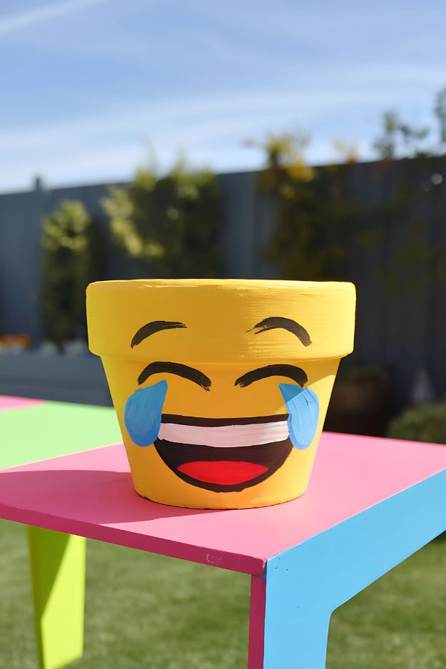 A yellow pot painted with a laughing emoji face, placed on a bright colored table outdoors.
