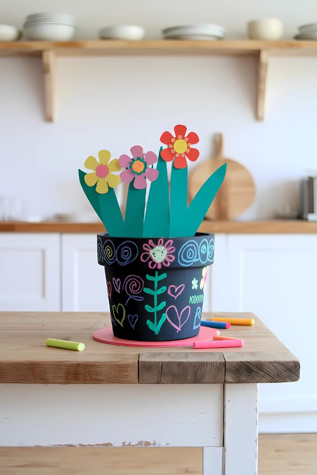 A flower pot with black chalkboard paint and decorated with colorful chalk doodles and paper flowers.