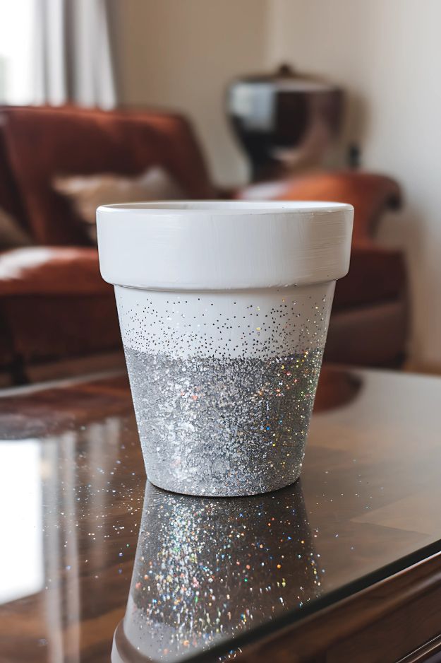 A white flower pot with its lower half dipped in silver glitter, sitting on a glass table.