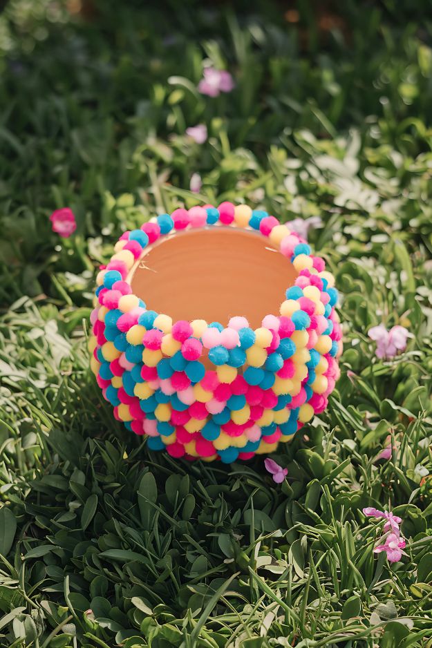A small pot completely covered in colorful mini pom-poms, sitting in the grass.