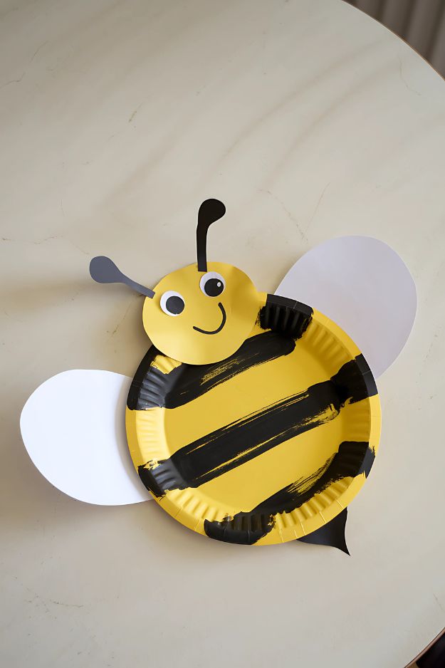 Smiling bee craft made from a yellow paper plate with black stripes and white wings.