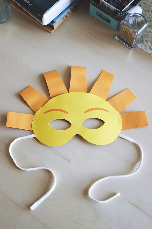 Yellow paper sunshine mask with orange rays and white string ties on a wooden table.