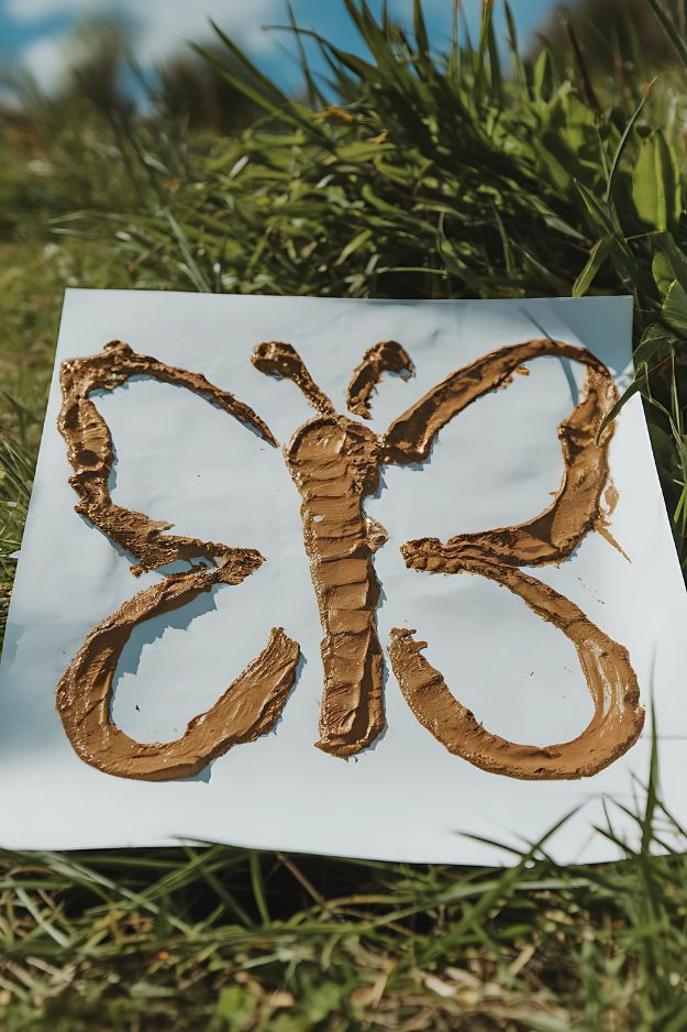 Mud-painted butterfly on white paper resting on grass in sunlight.