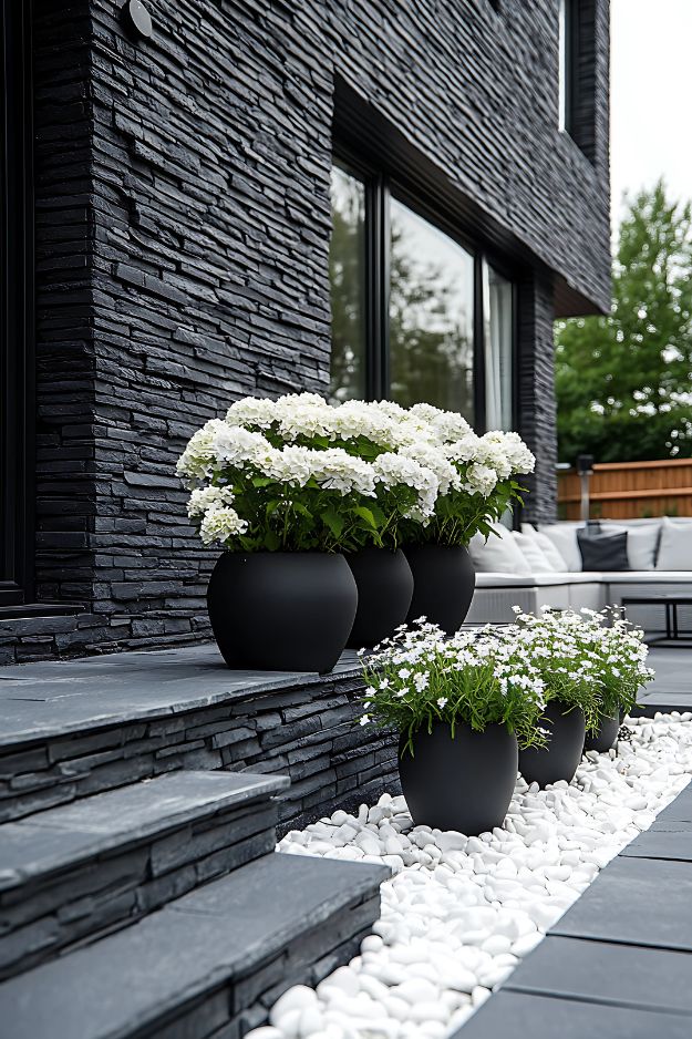 Black patio planters with white flowers and white rock border.