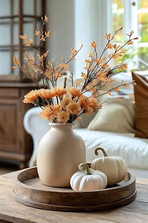 Round wood tray with beige vase of florals and two white pumpkins on coffee table.