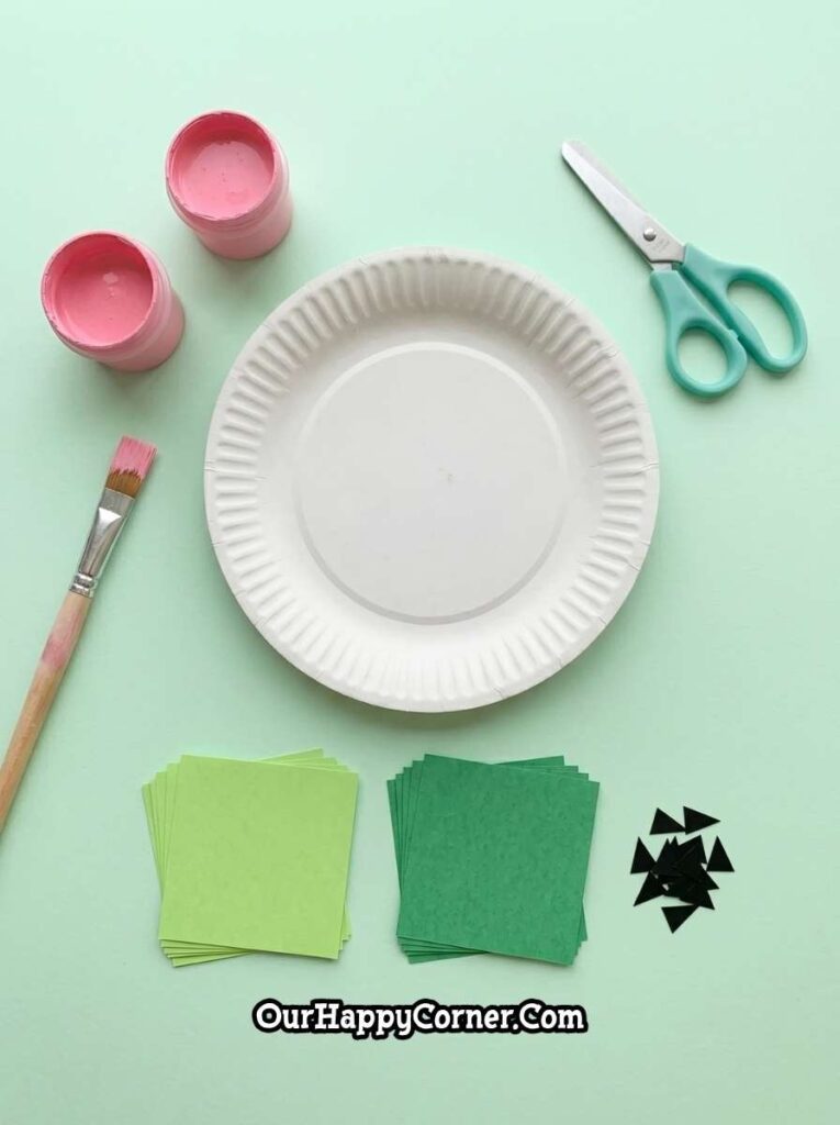 Materials including the paint, scissors, green paper squares, and black paper seeds on pastel background