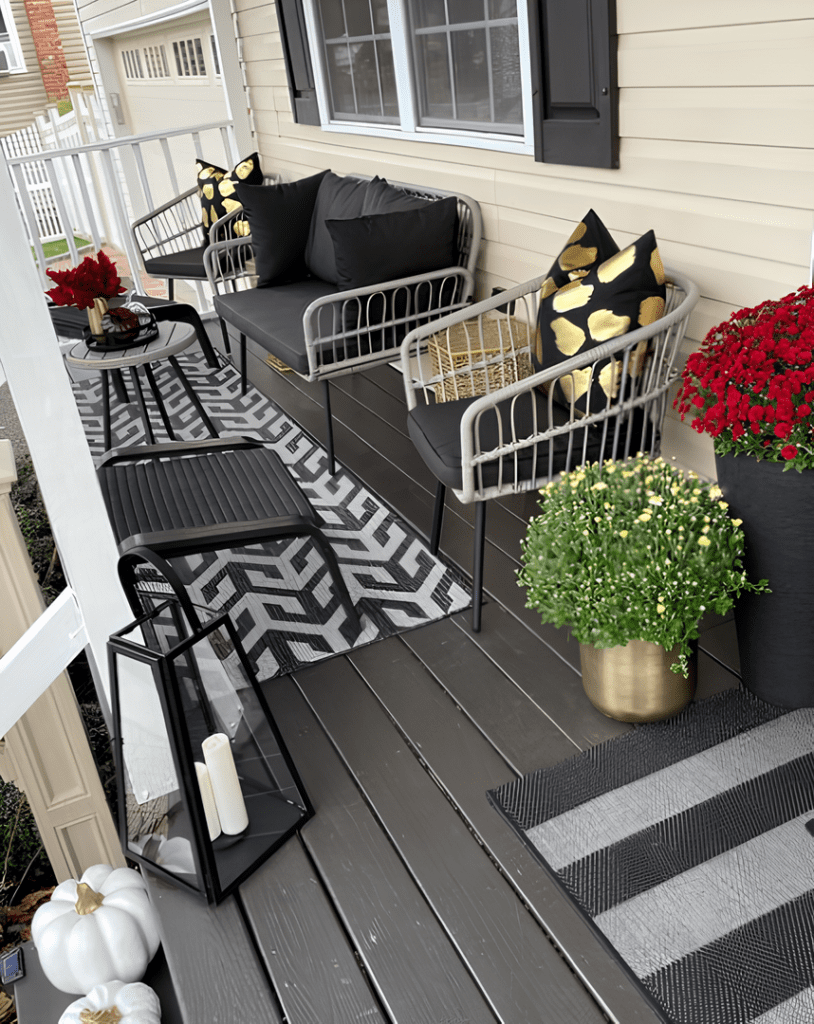 A porch decorated for fall with black and white geometric rugs, black cushioned chairs with gold and black pillows, potted mums, a black lantern with a candle, and white pumpkins.