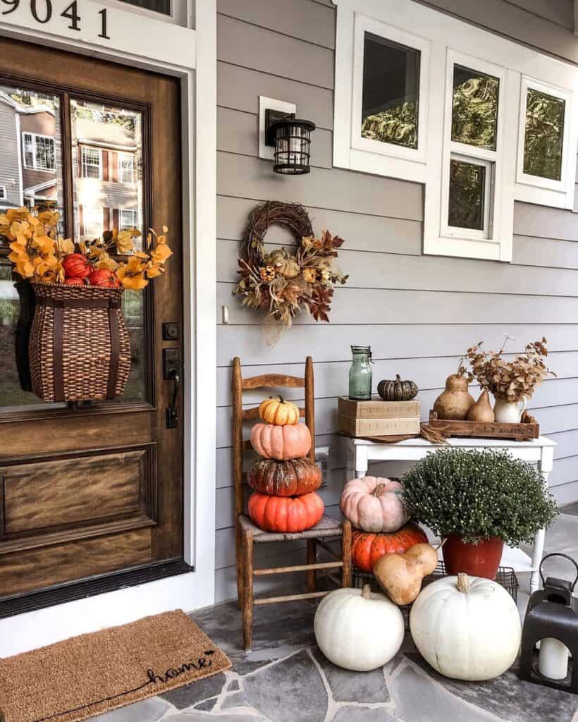 A cozy front porch decorated for fall with stacked pumpkins on a chair, a wicker basket filled with autumn foliage hanging on the door